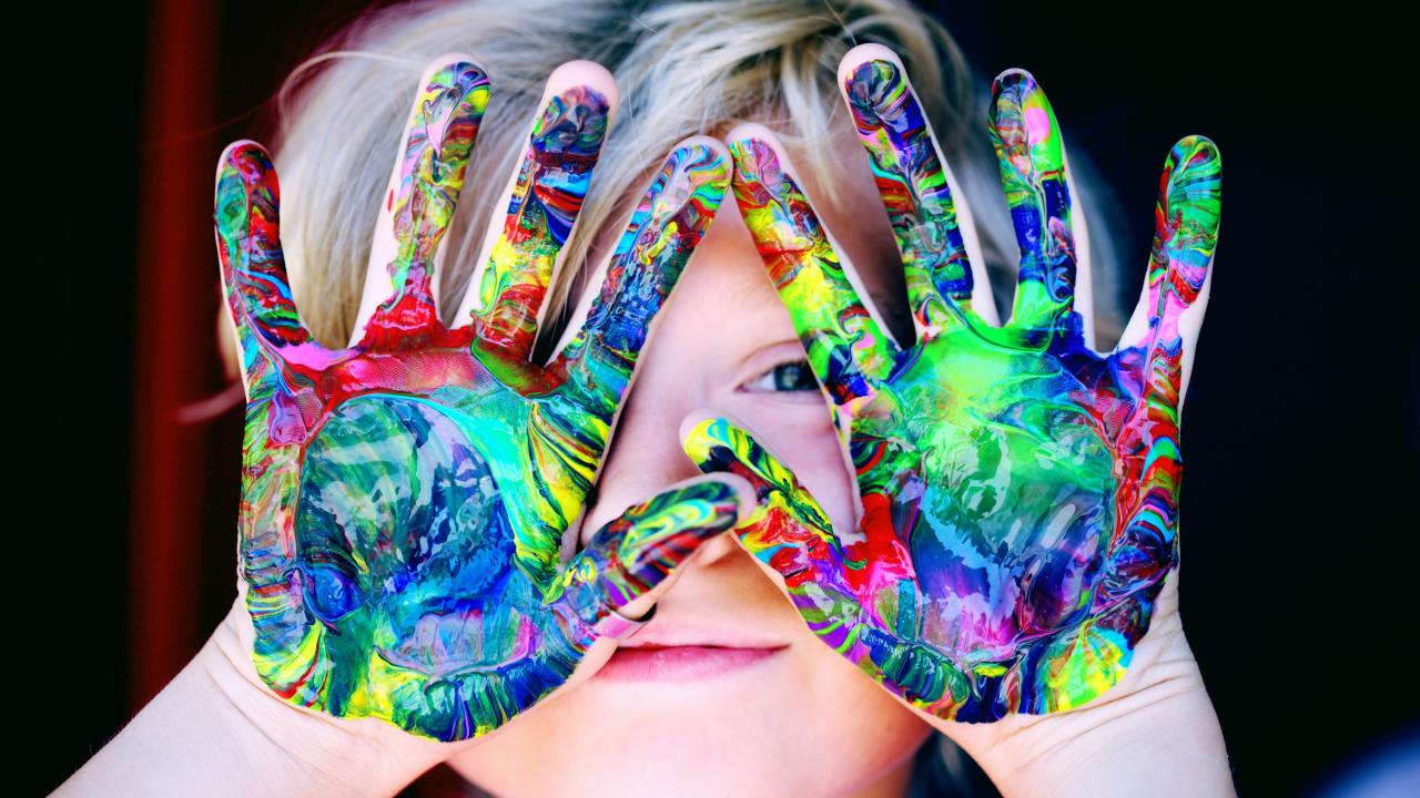 Child holds looks at the camera with paint-covered hands partially covering their face.